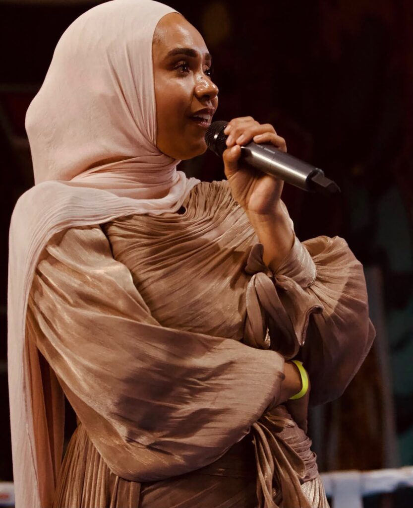 A profile of a woman with a headdress holding a mic to her mouth. She is wearing light pink and brown and the background is dark