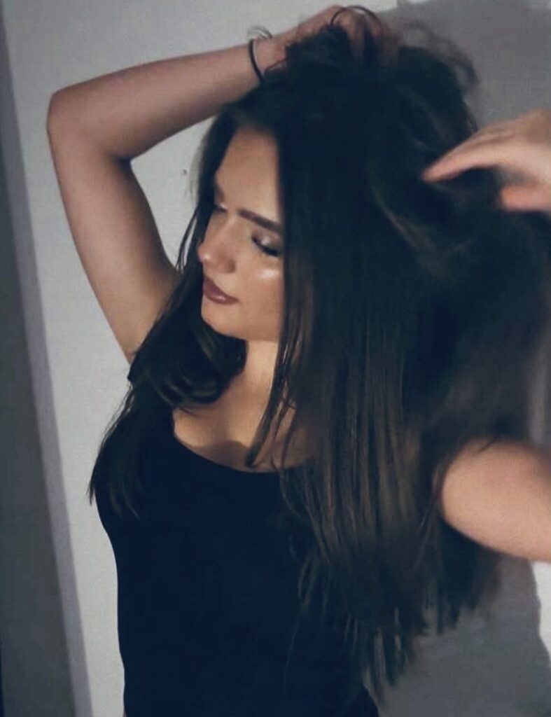 a young woman slightly to her side rubs her hands into the top of her head. She has long dark hair, a black vest and the background is white