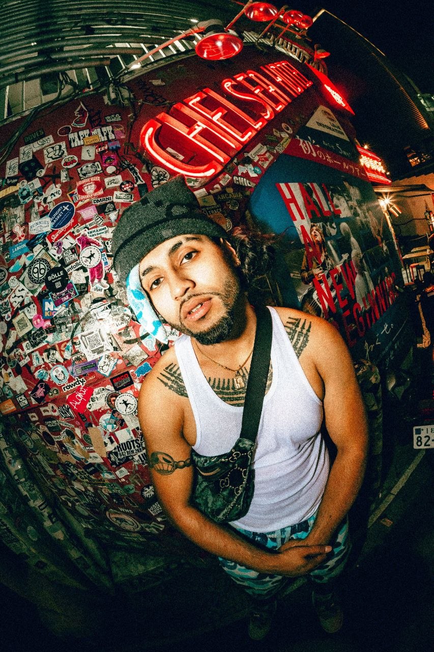 The camera looks down on a young man with a black had and white vest posing up to the camera with his hands held in front of him. Behind him is a background that's difficult to make out, perhaps lots of stickers and lights on a wall