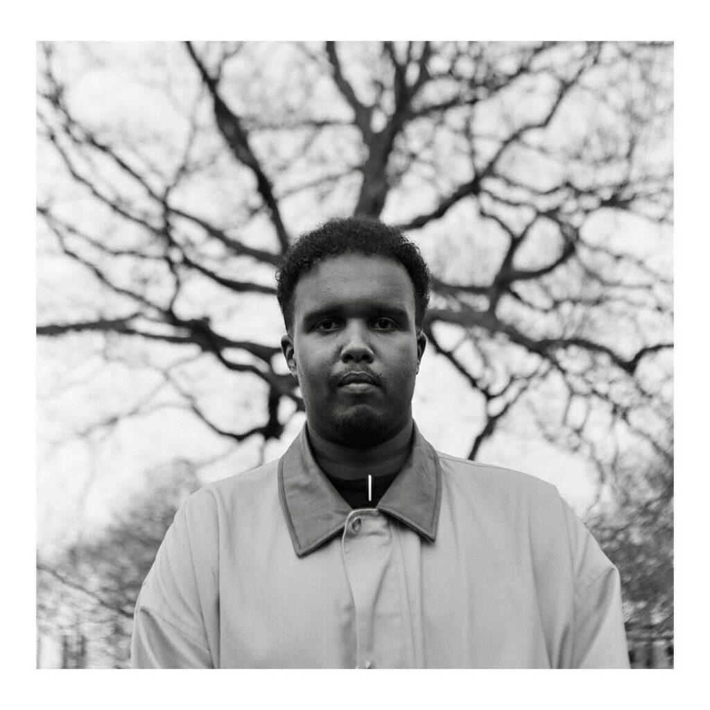 Black and white photo of a young man looking straight at the camera, wearing an anorak. In the background is a tree with wavy branches.